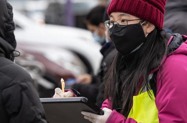 A masked safety worker writes on a clipboard in the street.