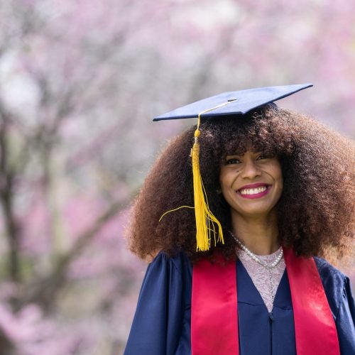 Black woman wearing graduation gown and cap with cherry blossom trees in the background