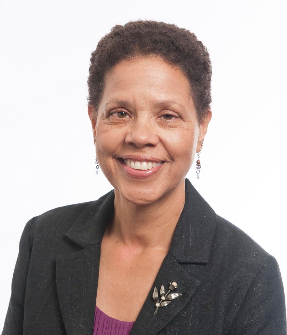 The photo features a smiling woman with short cropped hair on a white background. She is wearing a dark gray blazer with a fuchsia shirt underneath. She is wearing earrings and has a silver flower brooch on her left lapel.