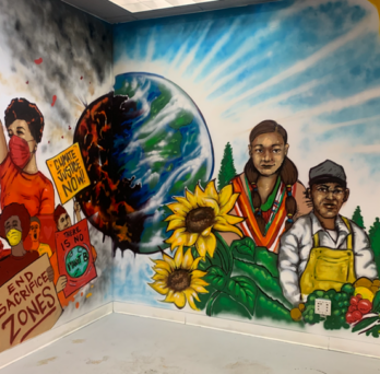 Environmental Justice Mural, featuring multiple people holding protest signs in front of a factory and gardeners in a field of pine trees and sunflowers. Between them Earth is split in two, one side is red, molten and cracked and the other is healthy and vibrant. 