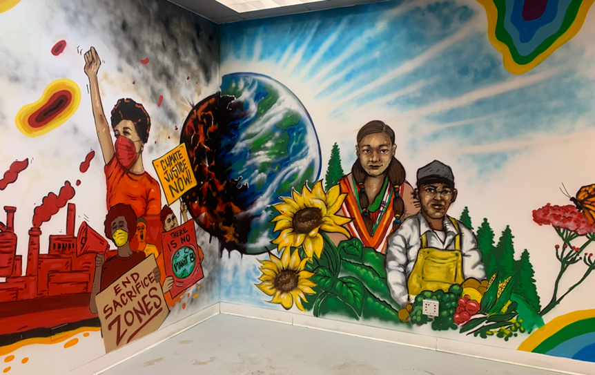 Environmental Justice Mural, featuring multiple people holding protest signs in front of a factory and gardeners in a field of pine trees and sunflowers. Between them Earth is split in two, one side is red, molten and cracked and the other is healthy and vibrant.