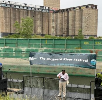 A single person stands beneath the banner of the Backward River Festival. The massive Damen Grain Silos tower in the background. 