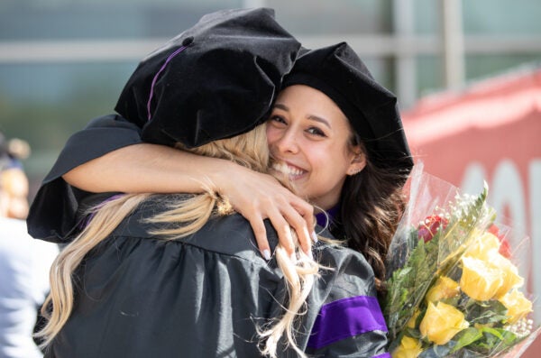 Two people in graduation gowns and hats hug with a bouquet of flowers.