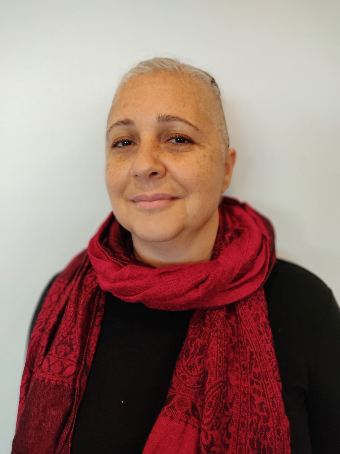 The photo features a person with short cropped white-grey hair standing against a cream-colored wall. She is wearing a black top with a maroon scarf in a paisley pattern looped around her neck, with the ends draped down her chest.