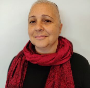 The photo features a person with short cropped white-grey hair standing against a cream-colored wall. She is wearing a black top with a maroon scarf in a paisley pattern looped around her neck, with the ends draped down her chest. 