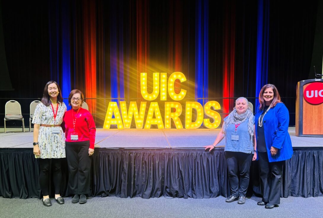 The photo features four people standing in front of an elevated stage. In the background on the stage is a large sign that says “UIC AWARDS” in all capital letters and is illuminated in gold with light bulbs inside of the letters. Behind the sign, the stage curtains are lit up with soft shades of alternating blue and red. Two of the people stand on the left side of the sign and two of the people stand on the right side, framing the sign between them.