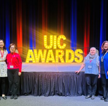 The photo features four people standing in front of an elevated stage. In the background on the stage is a large sign that says “UIC AWARDS” in all capital letters and is illuminated in gold with light bulbs inside of the letters. Behind the sign, the stage curtains are lit up with soft shades of alternating blue and red. Two of the people stand on the left side of the sign and two of the people stand on the right side, framing the sign between them. 