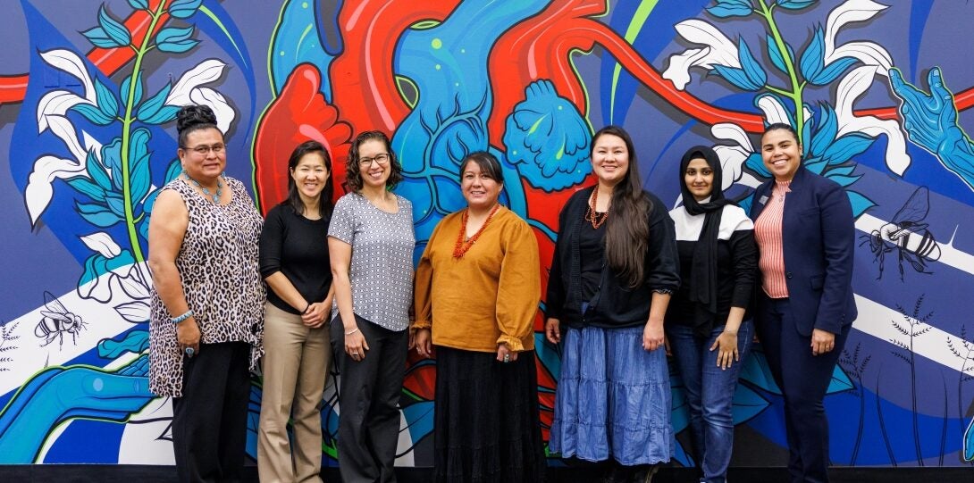 The photo features a row of seven people standing and smiling for the camera. Behind them is a large painted mural that fills the entire wall beyond the edges of the frame, featuring designs of hands, bees, blooming flower stalks, and grain in shades of blue, black and white. In the center of the mural is a large anatomical heart with red and blue chambers and arteries branching out into and connecting with the rest of the mural.