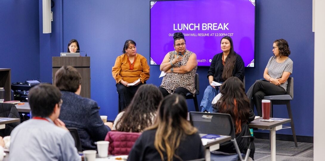 The photo features a panel of four people sitting on chairs in front of a large TV screen with a purple presentation slide. The second speaker from the left is talking into a hand-held microphone. To the left of the four seated people is one person standing at a podium, facilitating the panel. The walls of the room are blue, and in the foreground are people sitting at tables with their backs to the camera, listening to the presentation.