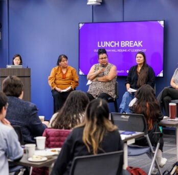 The photo features a panel of four people sitting on chairs in front of a large TV screen with a purple presentation slide. The second speaker from the left is talking into a hand-held microphone. To the left of the four seated people is one person standing at a podium, facilitating the panel. The walls of the room are blue, and in the foreground are people sitting at tables with their backs to the camera, listening to the presentation. 