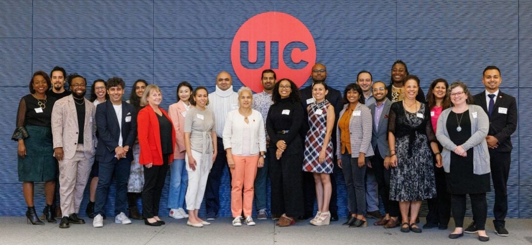 The photo features two rows of people, with 11 people standing in the back row and 12 people standing in the front row. They are standing against a blue textured wall with a large red circle in the center and the outline of the letters “UIC” cut out in the middle of the red circle.