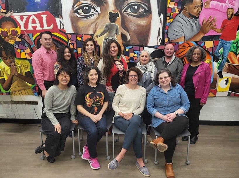 The photo features two rows of people--seven people standing with four people sitting in front of them on chairs. Behind them is a large painted mural that fills the entire wall beyond the edges of the frame, featuring various overlapping colorful scenes of joyful Black American life.