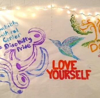 The photo features an art piece on a large white paper canvas with various painted designs and words. On the left side of the canvas, “Disability Cultural Center” is painted in bright blue with “= Disability Pride” painted below it in purple. Under these words are swirls like waves, leaves or petals in purple and grey-blue. In the center of the canvas is a side profile of a hummingbird with the feathers painted in bright blue, purple, blue-grey, and green. Under the hummingbird are the words “LOVE YOURSELF” in capital letters painted in red. On the right side of the canvas is the root, trunk and branch system of a tree painted in light brown with leaves and grass painted in green. Below the roots are the capital letters “DCC” painted in light brown, with some faint brown-red paint textured markings under it. The canvas is hung up on a white-painted brick wall and has white holiday lights strung along its upper edge and sides to frame it in a golden glow. 