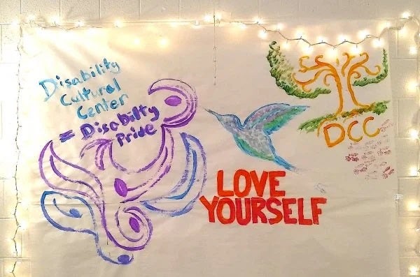 The photo features an art piece on a large white paper canvas with various painted designs and words. On the left side of the canvas, “Disability Cultural Center” is painted in bright blue with “= Disability Pride” painted below it in purple. Under these words are swirls like waves, leaves or petals in purple and grey-blue. In the center of the canvas is a side profile of a hummingbird with the feathers painted in bright blue, purple, blue-grey, and green. Under the hummingbird are the words “LOVE YOURSELF” in capital letters painted in red. On the right side of the canvas is the root, trunk and branch system of a tree painted in light brown with leaves and grass painted in green. Below the roots are the capital letters “DCC” painted in light brown, with some faint brown-red paint textured markings under it. The canvas is hung up on a white-painted brick wall and has white holiday lights strung along its upper edge and sides to frame it in a golden glow.