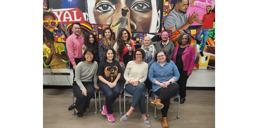 The photo features two rows of people--seven people standing with four people sitting in front of them on chairs. Behind them is a large painted mural that fills the entire wall beyond the edges of the frame, featuring various overlapping colorful scenes of joyful Black American life.