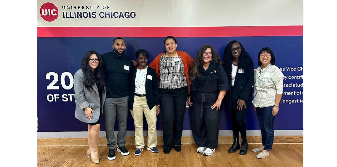 The photo features seven people standing and smiling against a wall with a large blue section listing facts about UIC behind them. There is a red band above the blue section and then a white section at the top, with the UIC logo in the far left corner.