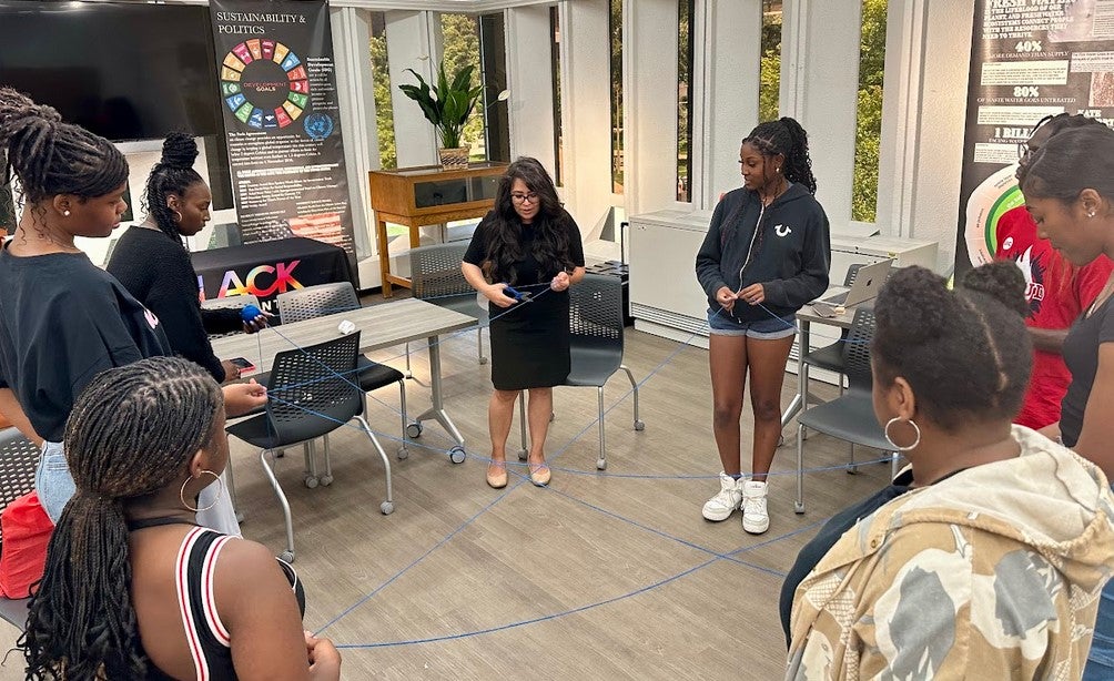 The photo features an instructor and several students standing in a circle in the middle of a UIC classroom. Each person is holding a part of a blue string and the string has many intersections as it runs between person to person. The instructor is holding a pair of scissors and preparing to cut the string. Posters about sustainability and politics as well as fresh water are propped up against the classroom walls.