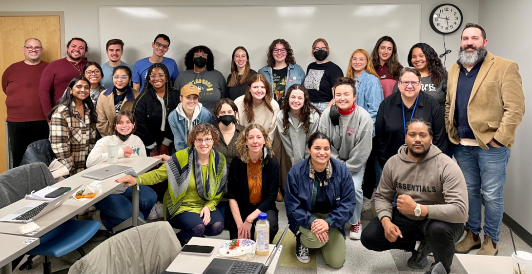 The photo features three rows of people--12 people standing, 10 people squatting, and five people kneeling in front. They are in an indoor classroom with a large white board behind them and tables with chairs on the left side of them.