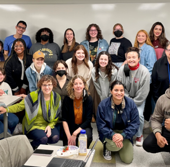 The photo features three rows of people--12 people standing, 10 people squatting, and five people kneeling in front. They are in an indoor classroom with a large white board behind them and tables with chairs on the left side of them. 