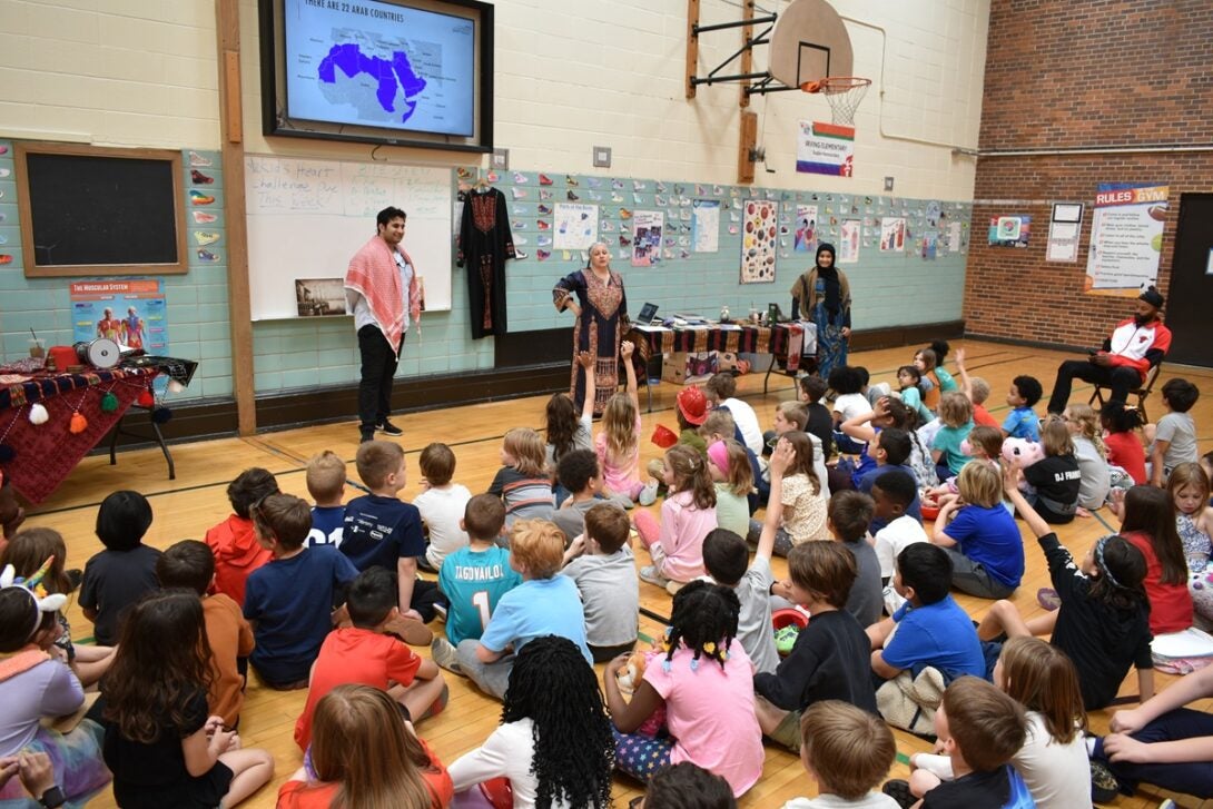 The photo features dozens of elementary school students sitting on a school gymnasium floor watching three university student presenters in traditional clothing from the Middle East and North Africa (MENA) regions. There are colorful tables set up behind the presenters with MENA regional artifacts and additional regional clothing hung up on the gymnasium wall. Above the presenters on the gymnasium wall is a projector screen with a PowerPoint slide showing the MENA region highlighted in purple.